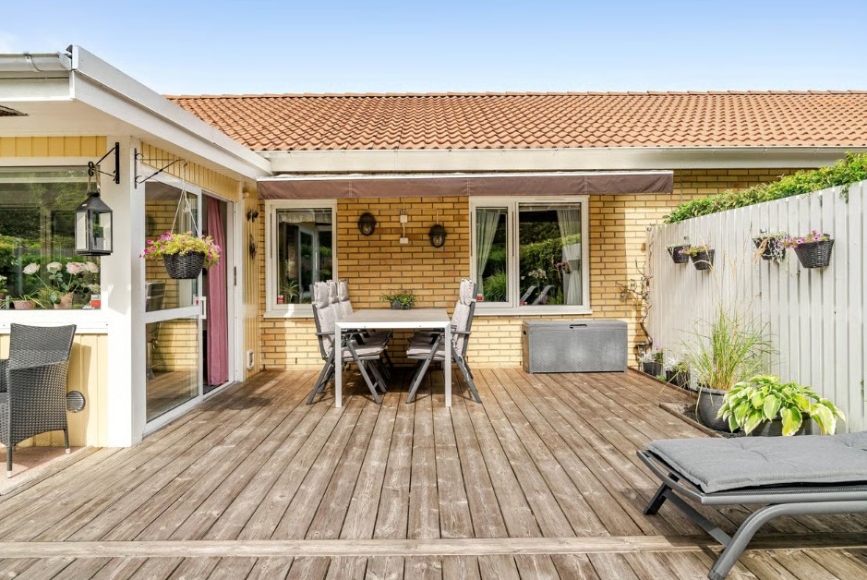 Uteplats på stort trädäck på baksidan, bord och stolar som står under utfällbar markis. Uterum ingång. Fönster från båda sovrummen mot baksidan.