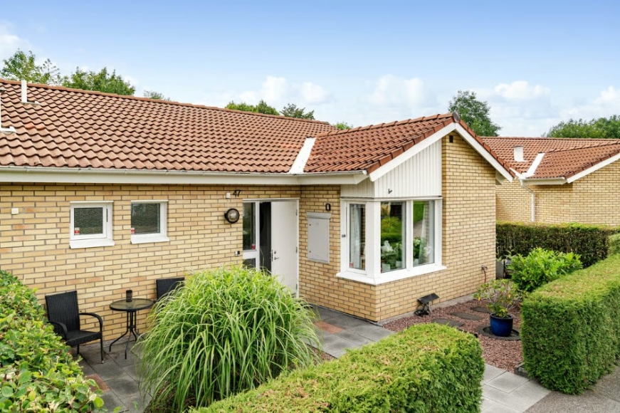 Framsida och entré till radhuset. Liten uteplats med bord och två stolar där man man njuta av morgonkaffet och frukost i solen. Kök och badrum åt entrén med utsikt över boulebanor och bostadsrättsföreningens lilla lekplats.